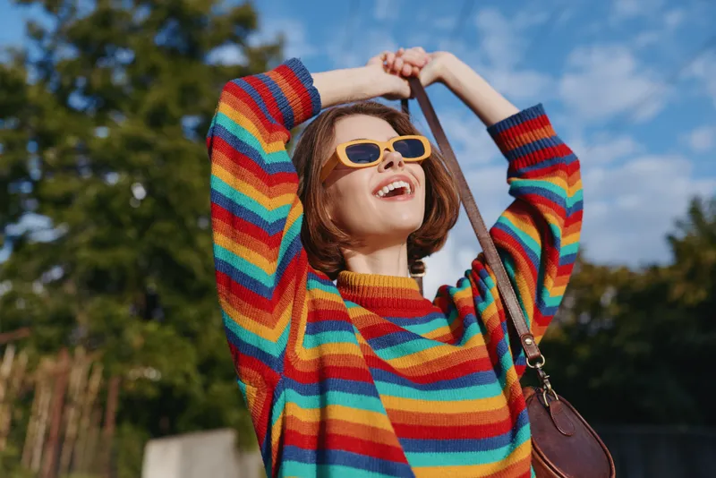 Young woman in a colorful striped sweater smiling with yellow sunglasses and casual outfit outdoors, summer scene with short hair and brown bag raising strap in joyful carefree pose.
