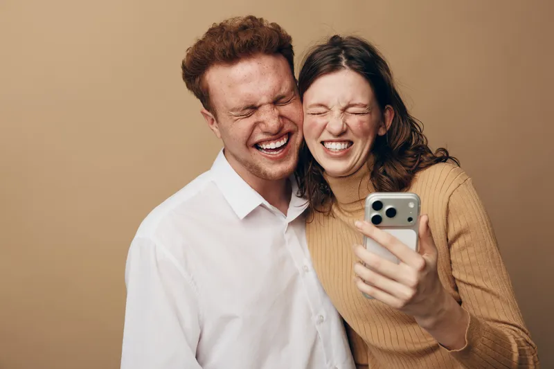 Happy young couple laughing together while taking a selfie with smartphone. Casual clothing and warm beige background create a joyful and intimate moment of friendship and fun.