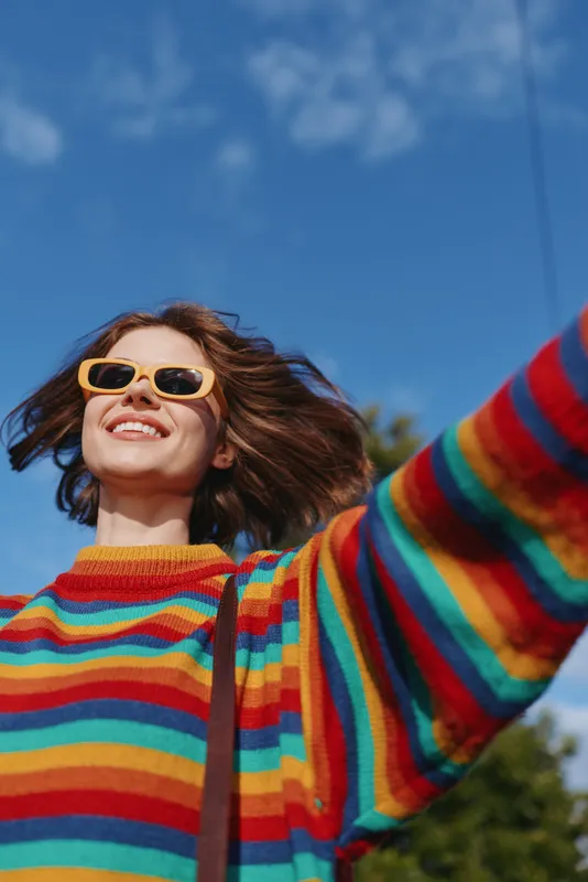 Woman smiling in a rainbow striped sweater with yellow sunglasses, mid 20s casual outfit, arms out and hair wind, joyful outdoors lifestyle under blue sky for travel and fashion