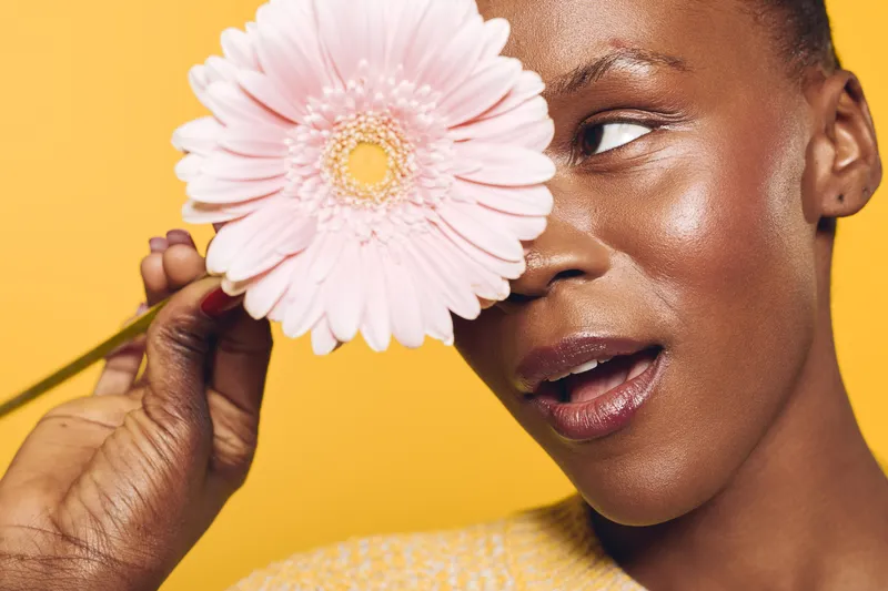 Close-up of a happy black woman with dark skin holding a pink flower near her eye on yellow background. Concept of beauty, skincare, natural glow, and floral wellness.