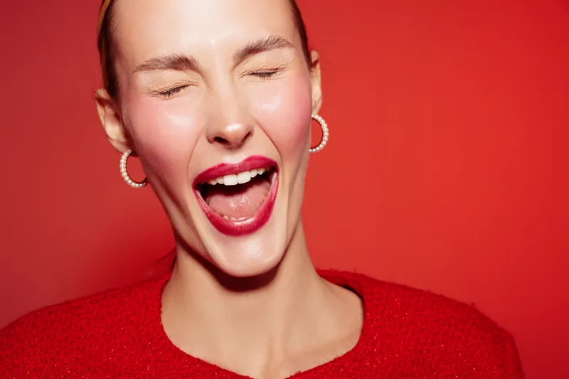 Close-up of a happy woman with closed eyes and open mouth laughing joyfully. She wears red lipstick and a red sweater against a red background. Concept of happiness and positive emotions.