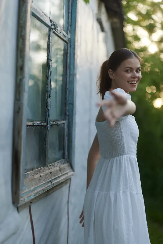 Young woman in a white dress reaching out, smiling, in a serene outdoor setting with a vintage window, evoking feelings of warmth and joy during spring