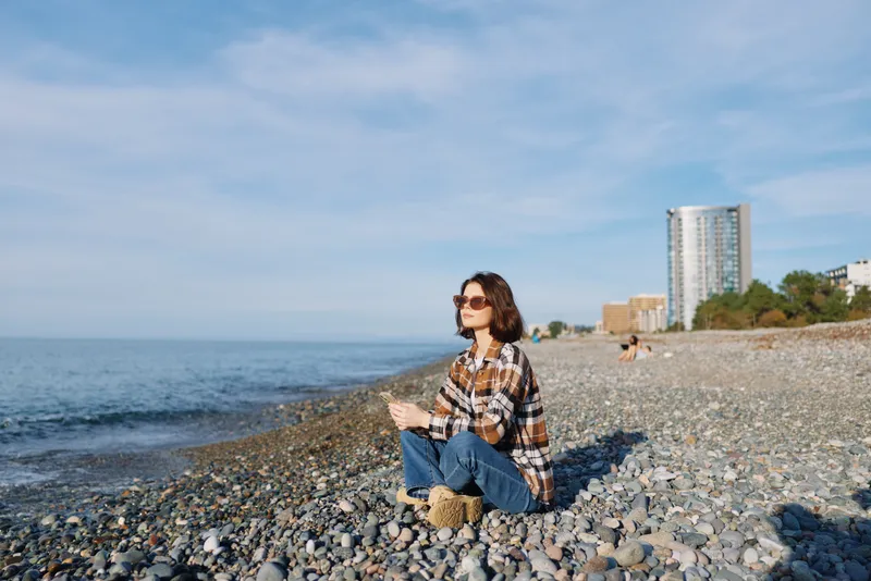 Beach scene with a woman sitting on a pebble shore, wearing a plaid shirt and jeans, sunglasses, enjoying quiet coastal view as city buildings rise in the distance.