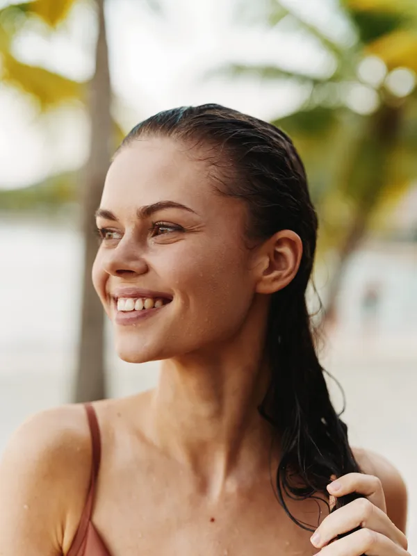 Smiling woman with wet hair enjoys a sunny tropical background, radiating happiness and warmth in a casual summer setting near palm trees.