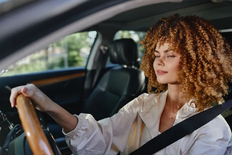 woman driving a car with curly hair and light jacket, wearing seatbelt, focused expression, modern interior, daytime natural light