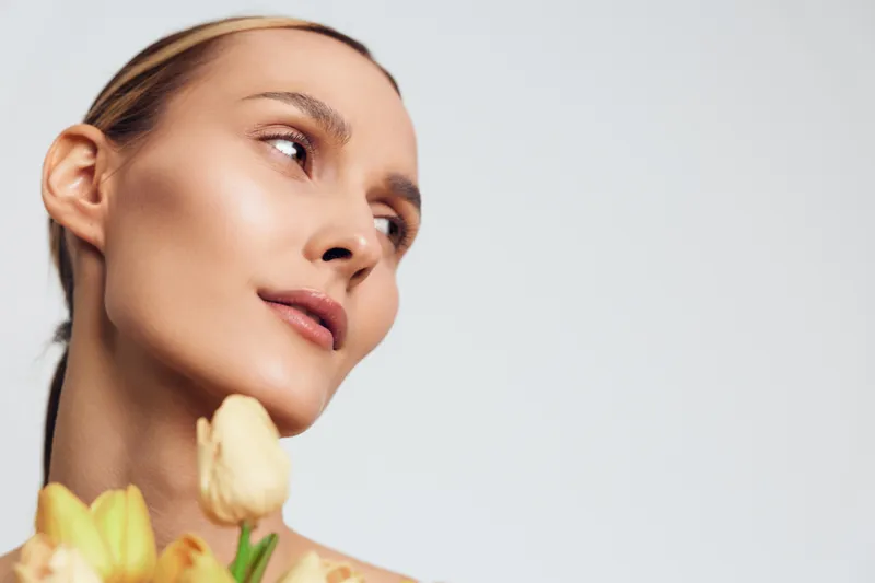 Portrait of a young woman with smooth skin looking to the side, holding yellow tulips, minimalistic background, soft natural light, elegant and fresh mood.