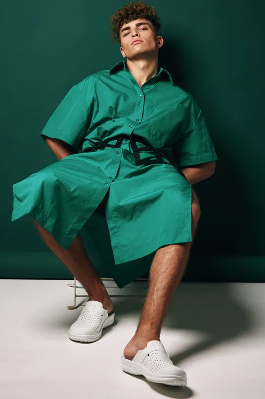 Young man wearing oversized green shirt dress with black belt and white slip-on shoes sitting on stool against dark green background in studio fashion portrait.