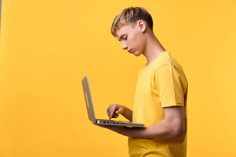 Young user in a yellow tee types on a laptop, focused on the screen against a vivid yellow background, depicting modern tech work, remote study, and casual computer usage in a bright setting
