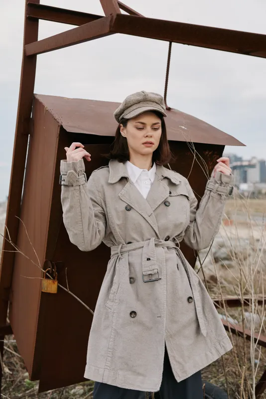 Young woman in gray checkered trench coat and matching cap stands outdoors near rusty metal structure, eyes closed. Fashion portrait in urban environment with muted colors and overcast sky.