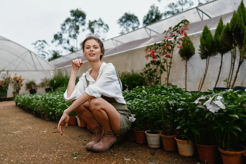 Young woman in a greenhouse, dressed casually, smiling among potted plants, showcasing a love for gardening in a natural, vibrant environment