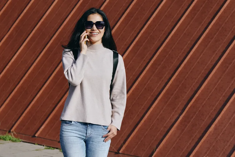 Woman on casual streetwear talking on phone, wearing sunglasses and jeans, standing beside a diagonal wooden fence, capturing outdoor lifestyle, fashion, confidence, and contemporary communication.