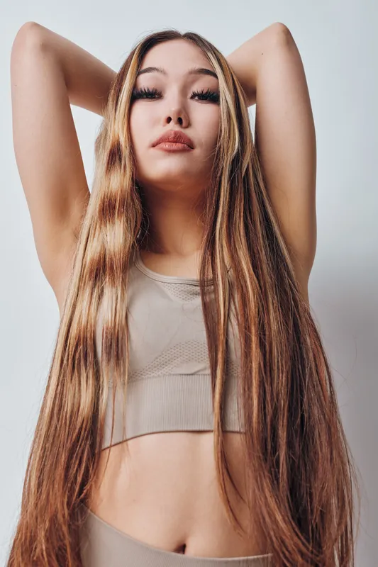 Young woman with long hair posing with arms raised wearing beige crop top and leggings, isolated on light background with confident expression and natural makeup.