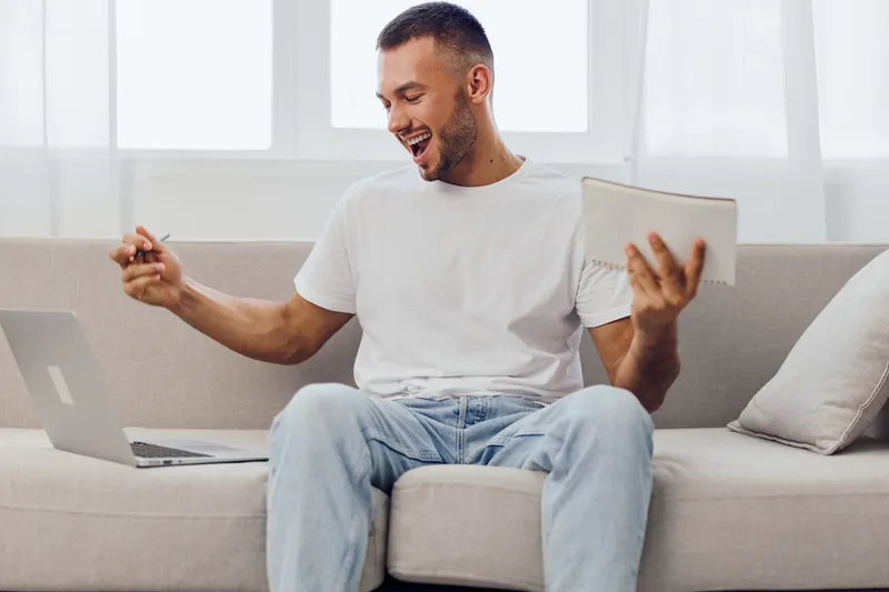 Excited man working on a laptop, holding notes and celebrating success. Bright, modern living room setting creates a positive and motivating atmosphere.