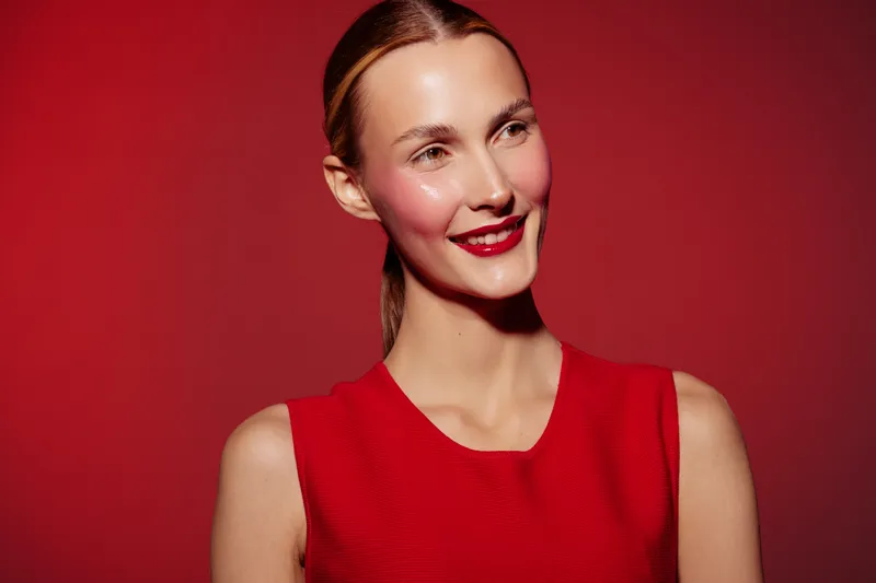 Smiling woman with red lipstick and red sleeveless dress poses against red background. Studio portrait with bright makeup and natural skin glow. Concept of beauty, fashion, and confidence.
