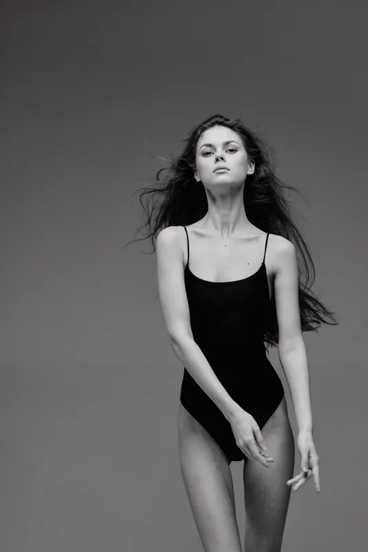 Black and white portrait of a confident woman wearing a black bodysuit posing against a gray background.