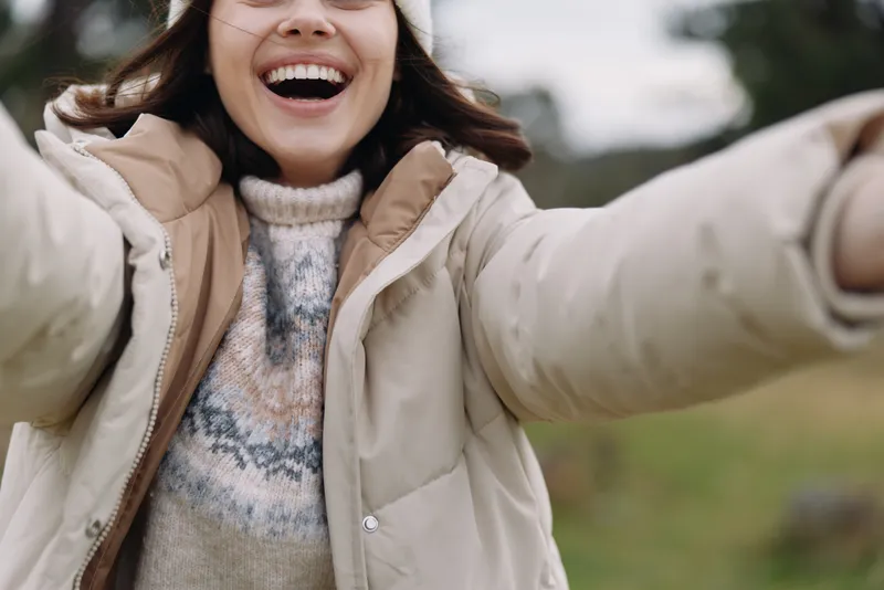 Outdoor portrait of a smiling woman in a warm jacket, taking a candid selfie in a park with a cheerful mood and natural daylight.
