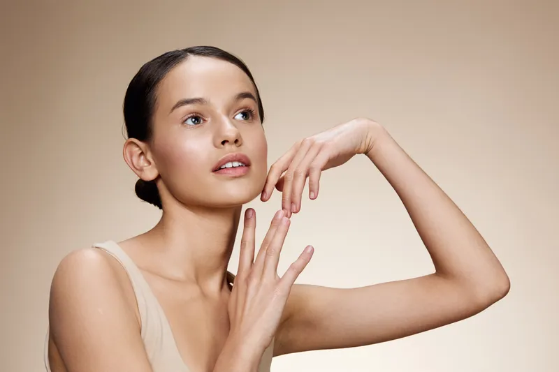 Elegant young woman with natural makeup and nude attire, showcasing a soft expression and graceful hands against a light beige background
