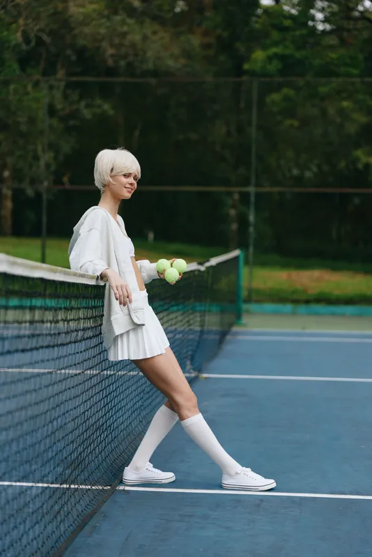 Young athletic woman with short white hair holding tennis balls stands confidently by the net on a sunny tennis court, dressed in a sporty outfit