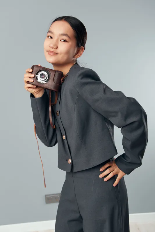 Young woman with short dark hair holding vintage camera smiling and posing in stylish gray suit on light background in studio. Portrait fashion concept with retro camera at casual outfit