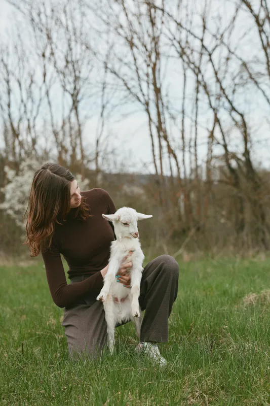 Woman in a brown top sits on grass, cradling a goat kid in an open field, smiling softly. Outdoor lifestyle scene highlighting nature, bonding, and gentle countryside calm, warmth, and connection.
