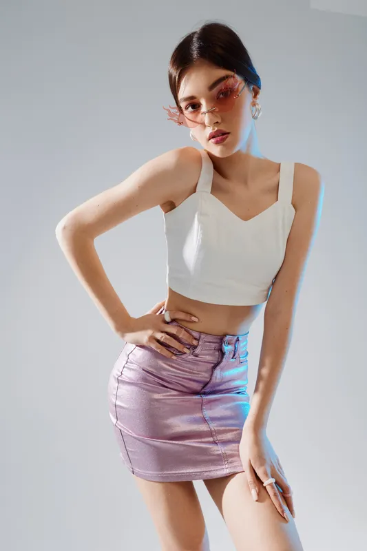 Young woman in a white crop top and shiny purple skirt poses confidently with hand on hip. She wears stylish pink sunglasses. Studio shot with soft lighting. Concept of fashion beauty.