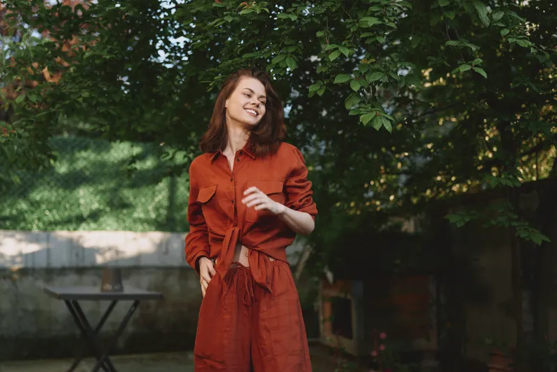 Smiling woman in a rust orange dress standing in a sunlit garden, casual pose and breeze in hair. Lifestyle portrait focusing on outdoor fashion and well being