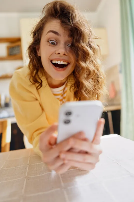 Excited young woman with curly hair takes a playful selfie indoors, leaning over a table with a bright smile, a cozy kitchen background, and warm natural light.