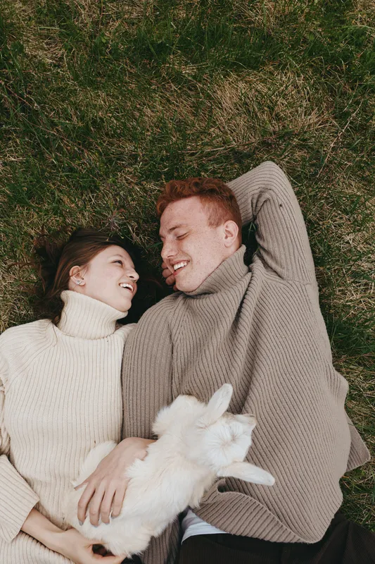 Cheerful couple outdoors, lying on grass with a goat kid, cozy sweaters, sunlit moment, relaxed pose, natural colors, rustic mood, countryside vibe