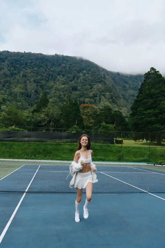 Young woman enjoying a refreshing drink on a tennis court surrounded by lush green mountains, exuding joy and vitality, perfect for healthy lifestyle concepts