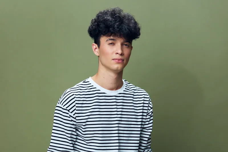 young man with curly hair wearing striped shirt posing against olive green background, casual style, relaxed expression, studio portrait lighting