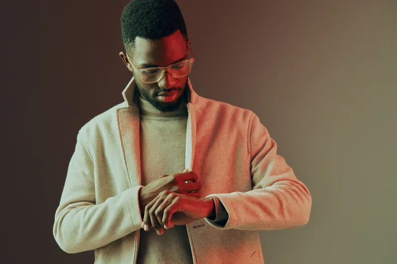Stylish man adjusting his watch, wearing a beige coat and sweater, exuding confidence against a warm gradient background