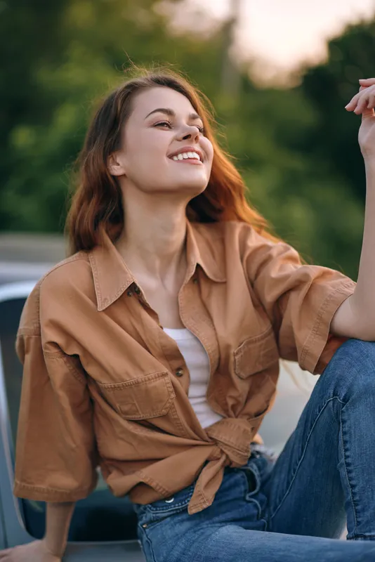 Young woman in casual brown shirt and jeans joyfully enjoying nature, with a bright smile, soft sunlight background, representing happiness and freedom.