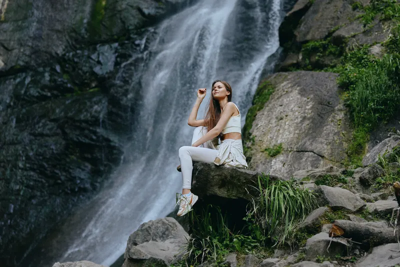Young woman sitting on rock near waterfall in casual white clothes, enjoying serene nature and peaceful outdoor environment with lush greenery around.