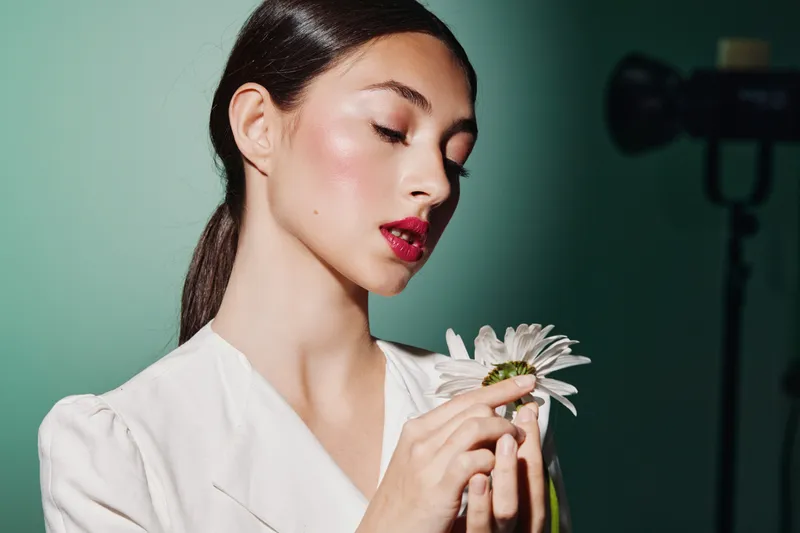 Elegant woman with perfect makeup gently holds light flower indoors on green backdrop. Concept emphasizes beauty portrait fashion creativity skin care and soft mood lighting.