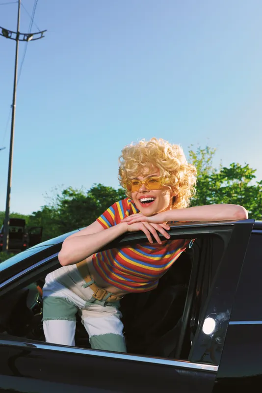 Smiling woman leaning out of a sleek car window, enjoying a sunny outdoors moment with a retro color palette. Emphasizes lifestyle, joy, and playful selfexpression.