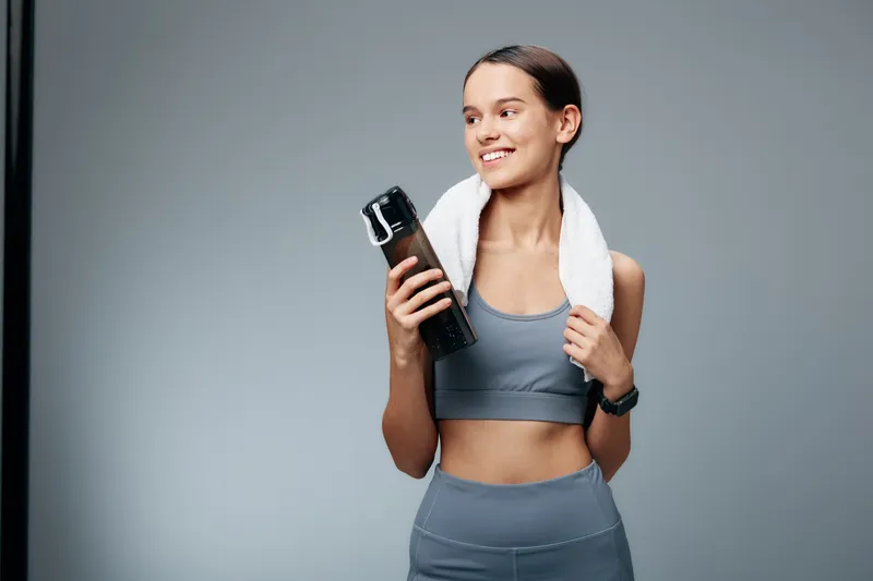 Smiling young woman fitness enthusiast in sportswear holds a water bottle and towel after workout, sporty pose and confident expression on a neutral studio background.