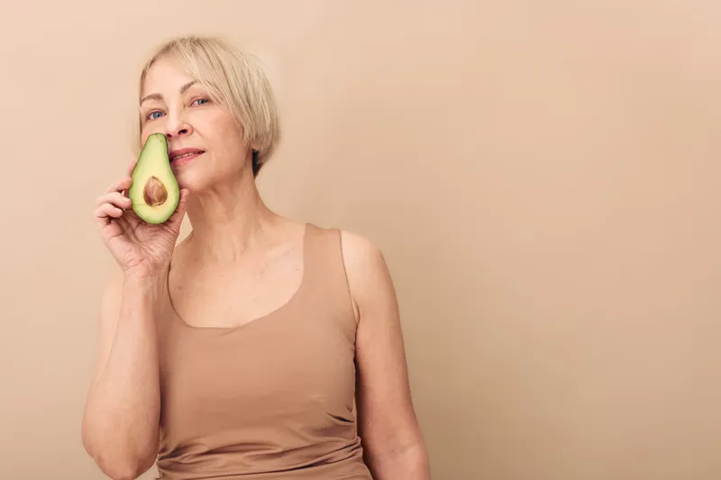 Mature woman with short blonde hair holds half of avocado near face, posing on beige background. Concept for healthy lifestyle, natural food, and skincare beauty.