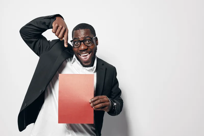 African American man with dark skin wearing glasses and black jacket smiling and pointing at red paper on white background for presentation or advertisement.