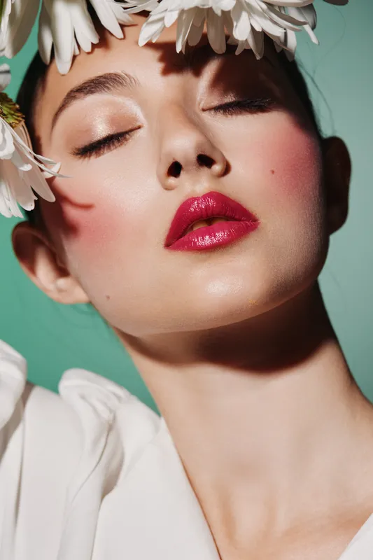 Close-up of a model with glossy red lips and flower petals on her head against mint background. Creative beauty fashion concept focused on makeup, softness, and natural freshness.