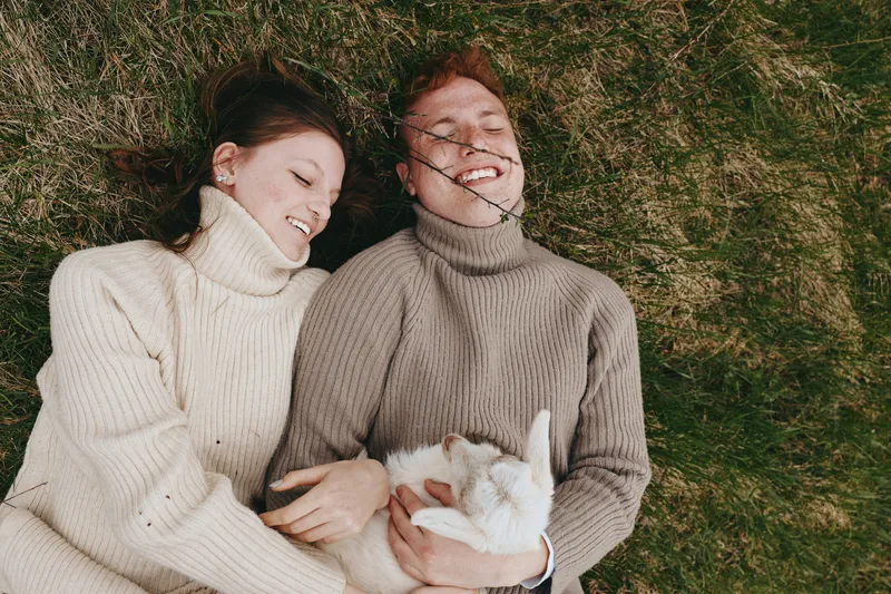 A young couple wearing cozy knitted sweaters lies on grass smiling happily while holding a small white baby goat, enjoying a relaxed moment outdoors in a casual lifestyle setting.