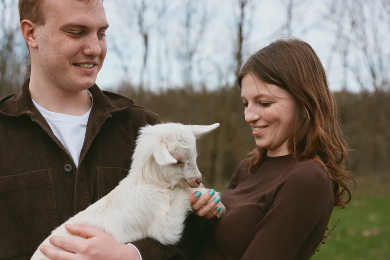 A young couple interacts with a small white baby goat outdoors during a casual lifestyle moment in early spring, smiling and enjoying the gentle animal together.