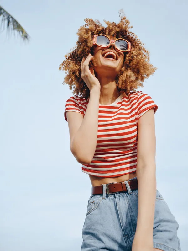 Vibrant portrait of a carefree young woman with curly hair, striped crop top, and sunglasses, captured against a bright blue sky and warm sunlight.