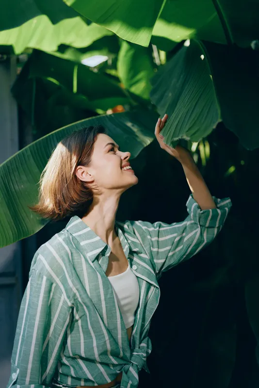 Young woman in a striped shirt interacts with large tropical leaves, enjoying a fresh, sunlit moment in lush greenery.