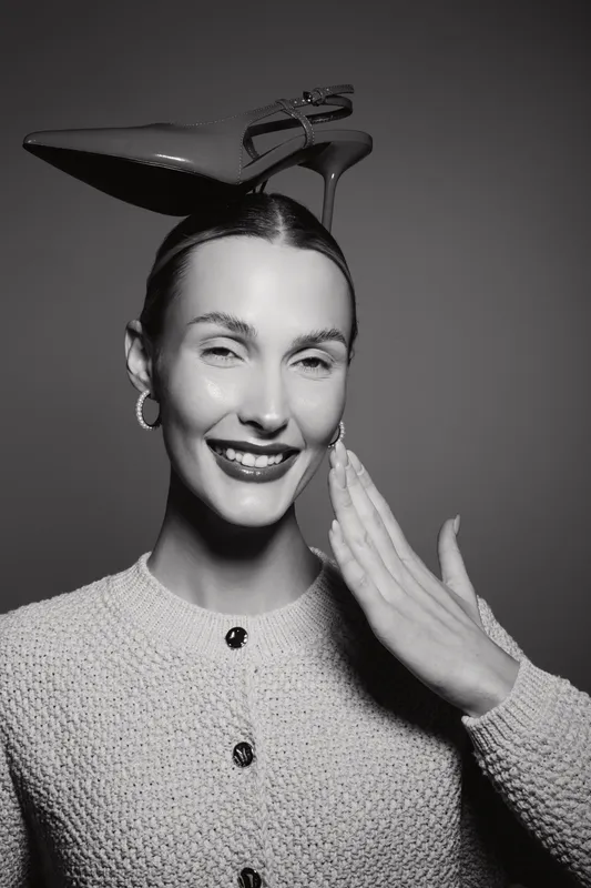 Caucasian woman with fair skin smiles while balancing a high heel shoe on her head, wearing a textured sweater and hoop earrings in studio lighting.
