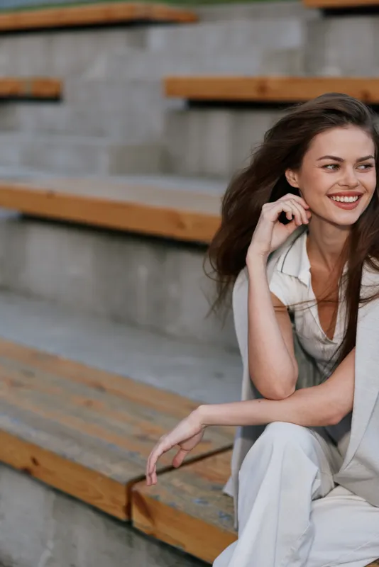 Smiling woman in light beige suit sitting on wooden bench with natural makeup and long hair, casual style, outdoor business fashion concept