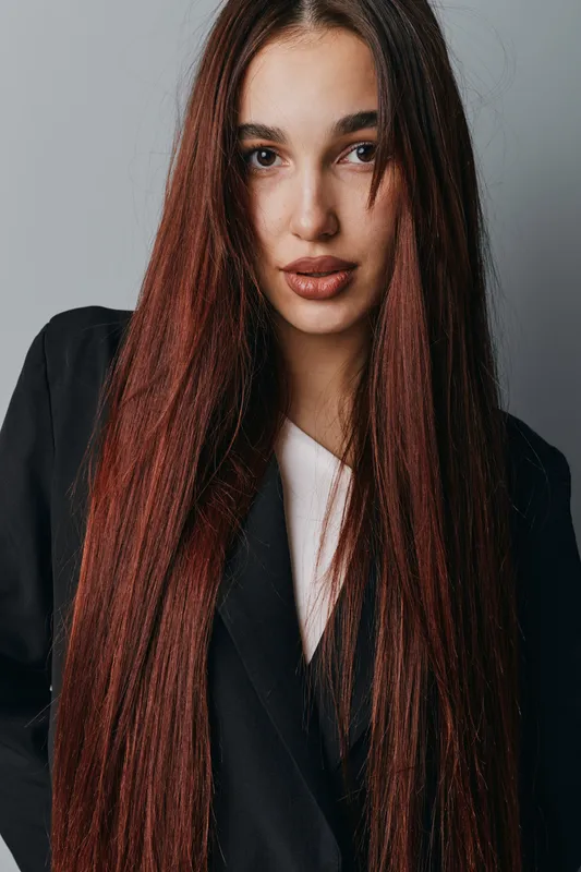 Young woman with long straight brown hair and braided strands wearing black blazer and white shirt posing in studio with neutral expression and natural makeup on face.