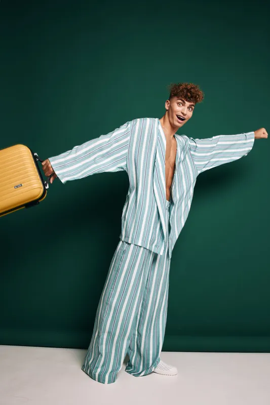 Young man in striped pajamas holding yellow suitcase with excited expression standing against green background. Casual travel, leisure, and vacation concept with comfortable clothing and luggage.