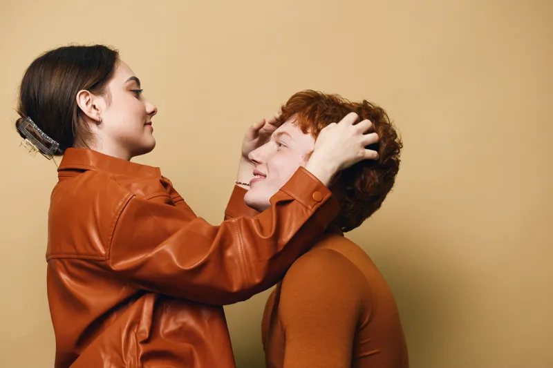 Woman and man in casual clothes smiling and touching each others heads against beige background. Loving relationship, friendship and happy people enjoying positive moment together.