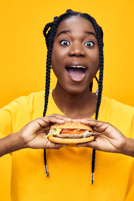 Excited girl holding a delicious burger on a vibrant yellow background, showcasing joy and enthusiasm, perfect for healthy eating concepts and fast food promotions