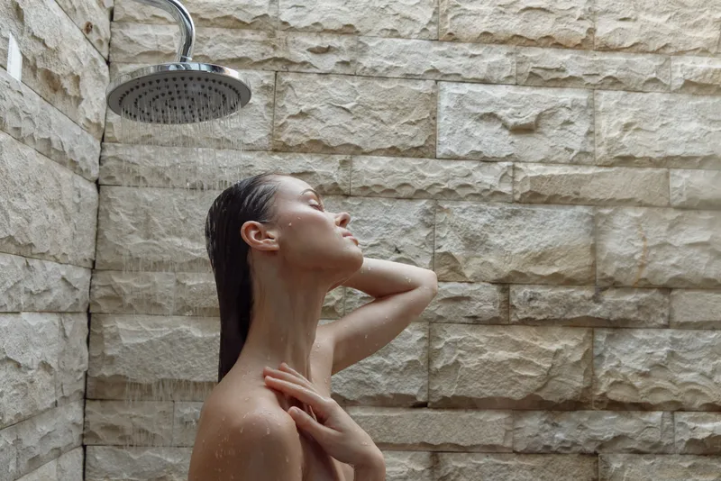 Woman enjoying outdoor shower with stone wall background, natural light, and water droplets falling, creating a serene and refreshing atmosphere in warm tones.